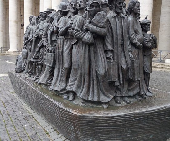 A bronze sculpture depicting a group of people standing closely together, each adorned in varied historical attire suggesting different eras and cultures, symbolizing migration or a journey. Their expressions are solemn and determined, with some holding suitcases and others carrying children.